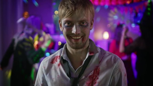 Halloween Costume Party: Portrait of Brain Dead Zombie Wearing Bloody Suit Smiles Creepily. In the Background Monsters Having Fun and Dancing in the Decorated Room