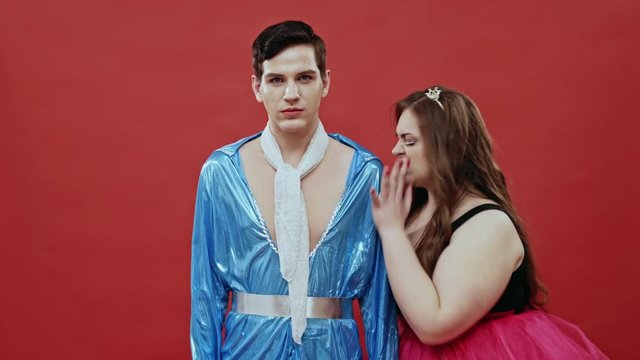 Overweight female in pink tutu dancing passionately around skinny guy in blue suit and grabbing him by balls in front of static camera on red background
