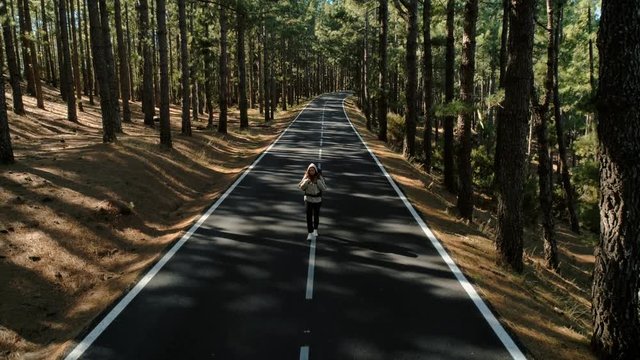 Aerial drone shot of young brave independent and strong woman during self discovering hiking, backpacking or hitchhiking trip through europe or usa walk in middle of empty epic forest road