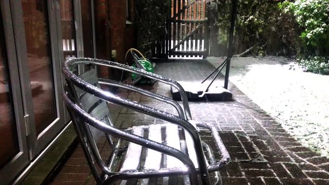 Snow falling on stacked metal chairs in backgarden with gate with chairs in centre of shot