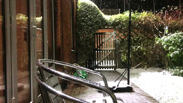 Snow falling on stacked metal chairs in backgarden in front of a wooden gate