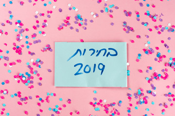 Hebrew text :"Elections 2019" on voting paper over pink with colorful confetti background. Israeli legislative Elections for the 21st Knesset Israel 9 April 2019.