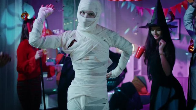 Halloween Costume Party: Old Skinny and Bandaged Mummy Dances. In the Background Zombie, Death, Witch and She Devil Have Fun in a Monster Party Decorated Room