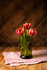 Simple rustic still life with blooming red tulips in a green glass on a wooden table with a napkin.