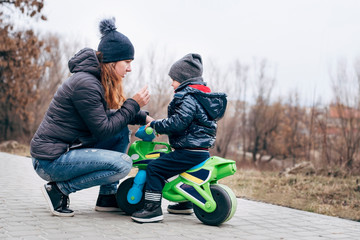 A woman is sitting near a little son sitting on a children's motorcycle. Mom explains son driving rules on the street