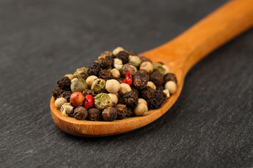 Macro shot of black, green, white and red peppercorn mix in a wooden spoon on a dark stone kitchen board. Shallow depth of field
