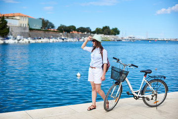 Obraz na płótnie Canvas Young slim woman standing near bicycle on stony sidewalk under clear blue sky on cozy harbor with sparkling water and cruise boat background. Tourism, active lifestyle and vacations concept.