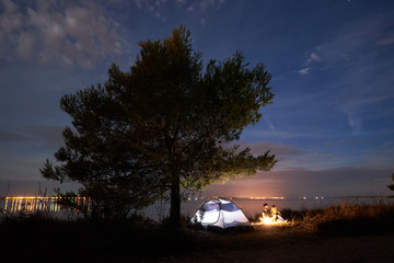Fototapeta na wymiar Tourist tent under big tree and young couple, bearded man and pretty woman hikers having a rest at night on sea shore by campfire under blue evening sky. Tourism, camping and loving relations concept.