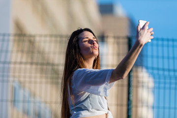Front view of beautiful trendy young woman wearing casual wear standing in the street while taking a selfie smiling in a sunny day