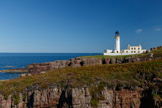 Rua Reidh lighthouse with keepers house on a rocky, grassy coastline with blue sky.