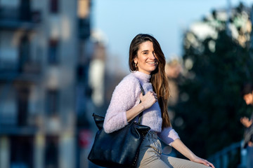 Front view of beautiful trendy young woman smiling and wearing casual wear sitting on a metallic fence in the street while looking camera in a sunny day