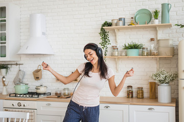 Young happy beautiful woman dancing in kitchen in pajamas and headphones, listening to music