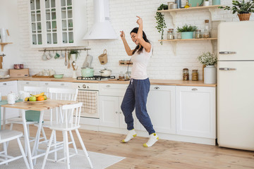 Young happy beautiful woman dancing in kitchen in pajamas and headphones, listening to music - 253833173