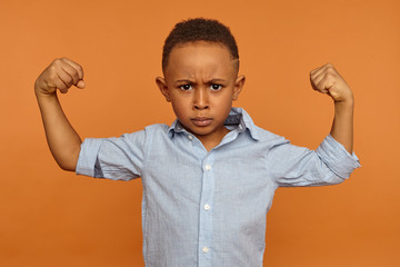 Serious seven year old dark skinned boy having grumpy facial expression, frowning and tensing biceps, demonstrating his strong arms, being full of energy. Strength, vitality, health and well being