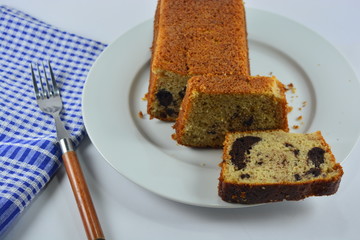 a banana cake on a white plate next to a checked napkin and a metal fork with a wooden handle on a white background