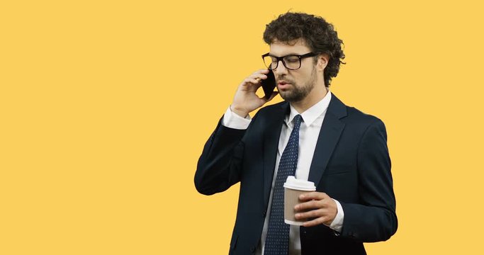 Attractive businessman in glasses and tie talking on his smartphone while drinking coffee to go on the yellow background.