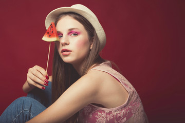 Attractive Lovely girl with pink make up, wearing jeans, hat and top, posing at red studio background, holding slice watermelon and eat it , emotionally look, close up portrait