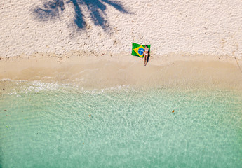 Aerial view of a young woman with a brazilian flag on the beach. Crystal clear water, tropical vacation scenario, coconut tree and boats. Drone view