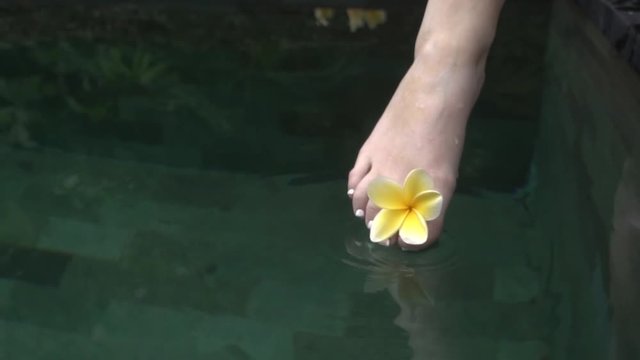 Young woman foot with a yellow flower touching the water surface.