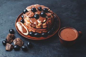 Hot chocolate drink in a cup and pancakes with banana, chocolate sauce and grapes in plate, on dark background