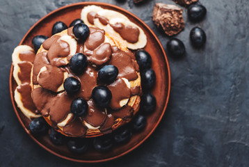 Pancakes with banana, chocolate sauce and grapes in plate, on dark background, top view