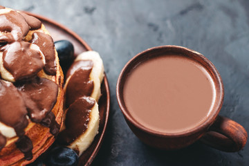 Hot chocolate drink in a cup and pancakes with banana, chocolate sauce and grapes in plate, on dark background