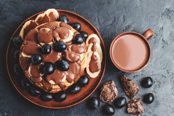 Hot chocolate drink in a cup and pancakes with banana, chocolate sauce and grapes in plate, on dark background, top view