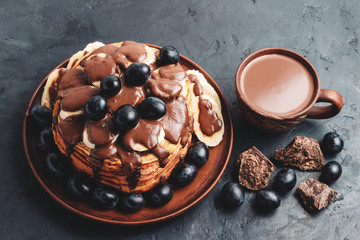 Pancakes with banana, chocolate sauce and grapes in a plate and hot chocolate drink in a cup on dark background
