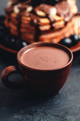 Hot chocolate drink in a cup and pancakes with banana, chocolate sauce and grapes in plate, on dark background