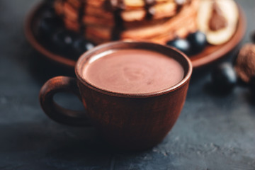 Hot chocolate drink in a cup and pancakes with banana, chocolate sauce and grapes in plate, on dark background