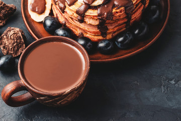 Hot chocolate drink in a cup and pancakes with banana, chocolate sauce and grapes in plate, on dark background