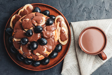 Hot chocolate drink in a cup and pancakes with banana, chocolate sauce and grapes in plate, on dark background, top view