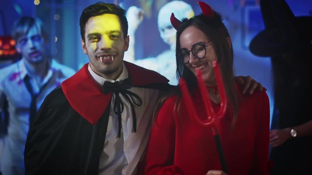 Halloween Costume Party: Portrait of Seductive Count Dracula Smiling and Hugging Dazzling and Bewitching She Devil with a Trident. In the Background Fun Monster Party with Disco Ball Light Show