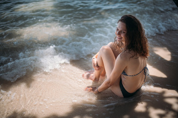 Young slim girl sitting in the shade of a tree in the sea waves on the coast of a tropical island