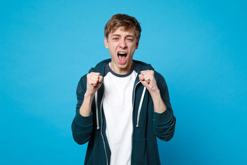 Portrait of unhappy young man in casual clothes screaming, clenching fists isolated on blue wall background in studio. People sincere emotions, lifestyle concept. Mock up copy space. Advertising area.