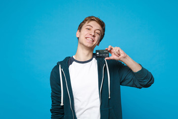 Fototapeta premium Portrait of cheerful young man in casual clothes holding credit bank card isolated on blue wall background in studio. People sincere emotions, lifestyle concept. Mock up copy space. Advertising area.