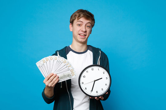 Cheerful Young Man In Casual Clothes Holding Fan Of Money In Dollar Banknotes, Cash Money, Round Clock Isolated On Blue Wall Background. People Sincere Emotions, Lifestyle Concept. Mock Up Copy Space.