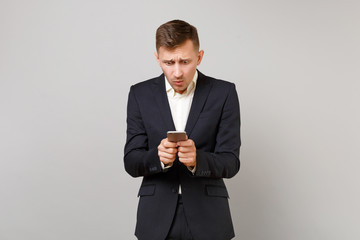 Shocked concerned young business man in classic black suit using mobile phone, typing sms message isolated on grey background in studio. Achievement career wealth business concept. Mock up copy space.