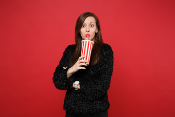 Portrait of amazed young woman in black fur sweater drinking cola or soda from plastic cup isolated on bright red background in studio. People sincere emotions, lifestyle concept. Mock up copy space.