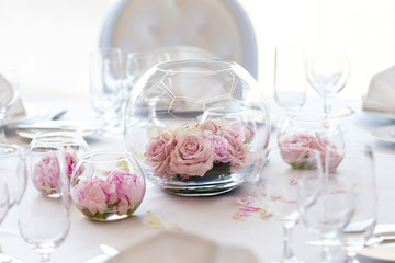 Detail of white wedding table set-up with fresh pink roses in glass bowls and rose petals. Blurry background.