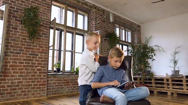 Cute little boy runs to his older brother who learning online English lessons on a computer tablet while sitting in an armchair in modern schoolroom. Indoors. Children education, science, technology