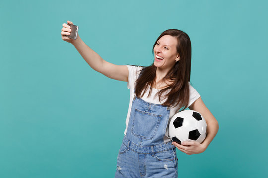 Smiling young woman football fan with soccer ball doing selfie shot on mobile phone isolated on blue turquoise background. People emotions, sport family leisure lifestyle concept. Mock up copy space.
