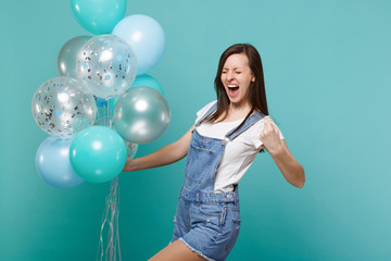 Happy screaming young woman with closed eyes doing winner gesture, celebrating and holding colorful air balloons isolated on blue turquoise background. Birthday holiday party, people emotions concept.