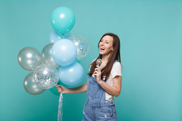 Fototapeta premium Cheerful young woman in denim clothes sing song in microphone, celebrating and holding colorful air balloons isolated on blue turquoise background. Birthday holiday party, people emotions concept.