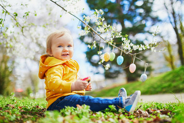 Cute little one year old girl playing egg hunt on Easter. Toddler sitting on the grass under apple tree in full bloom decorated with colorful eggs