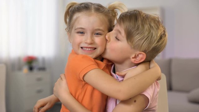 Younger sister hugging brother drawing at table, kissing cheeks each other, love