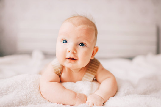 Adorable lovely newborn male baby boy with smiling emotional  happy face lifestyle indoor portrait. Funny infant child lying on stomach on bed with white wall on background.  Carefree childhood.