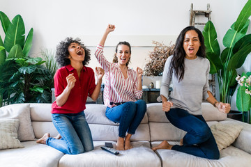Three excited women on couch at home watching Tv and cheering