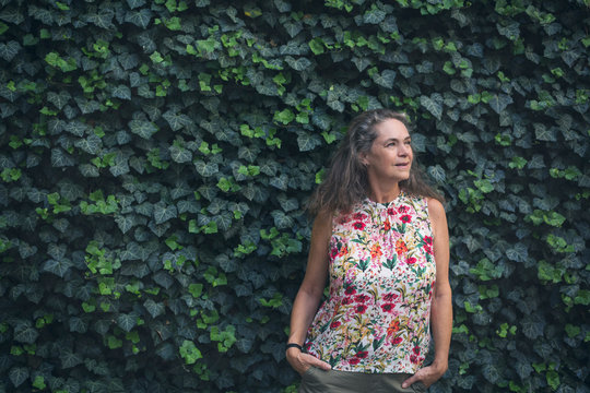 Portrait of relaxed mature woman standing in front of wall overgrown with ivy wearing top with floral design