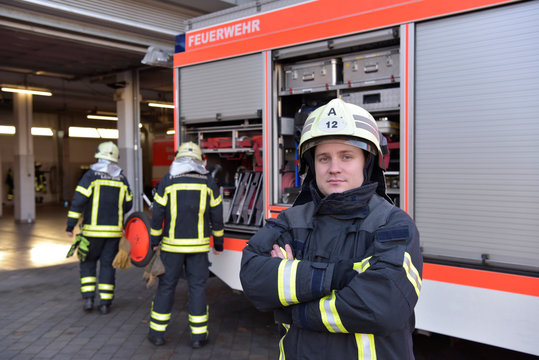 Portrait of confident firefighter in front of fire engine with colleagues in background